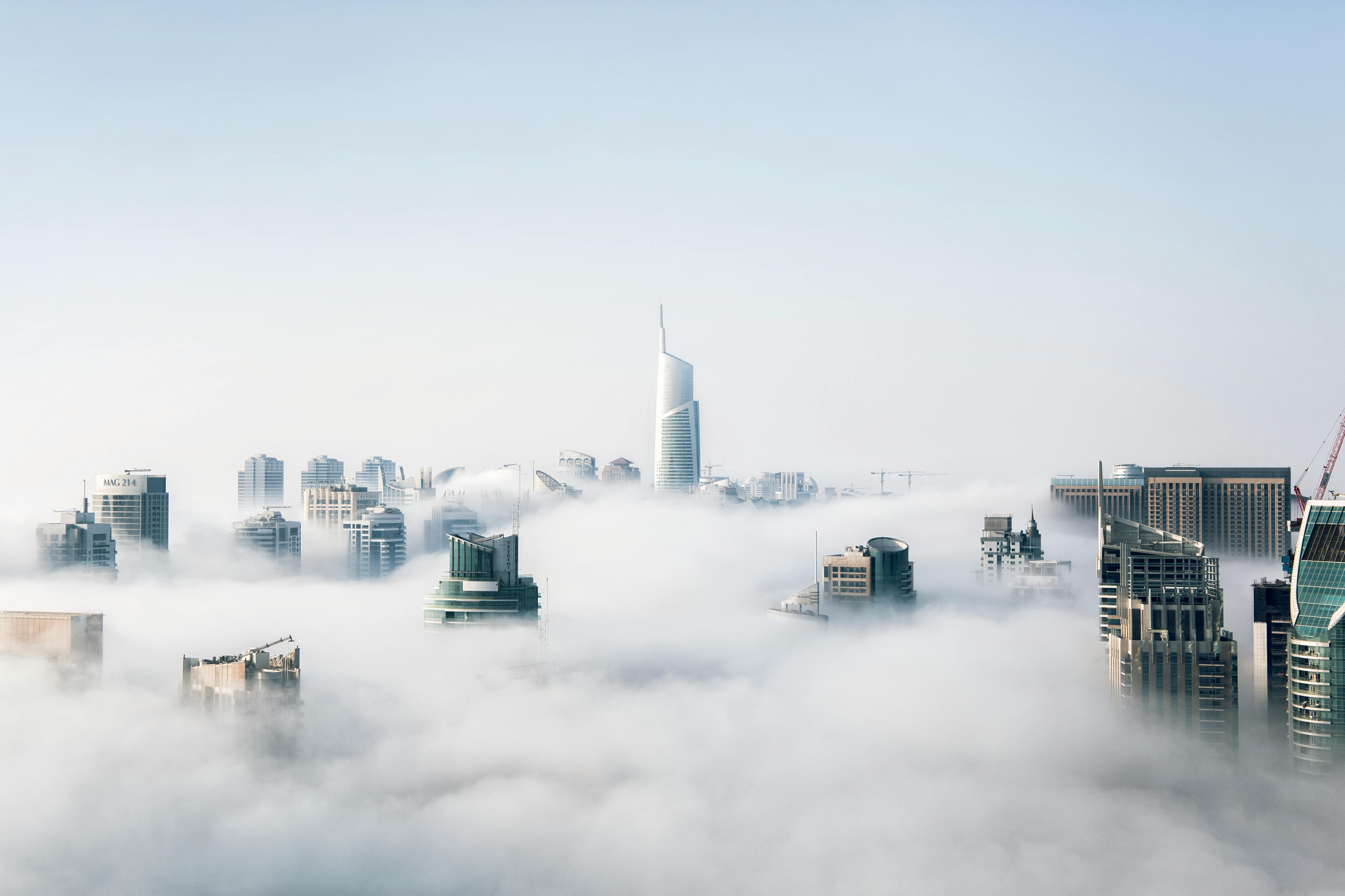 Skyline van een moderne stad gehuld in wolken, met alleen de toppen van wolkenkrabbers zichtbaar boven een dichte mistlaag. Een grote, opvallende toren steekt uit in het midden van de horizon. De scène symboliseert cloud computing.